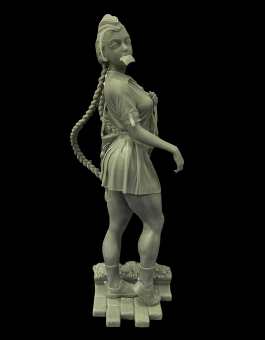 Unpainted 3D printed Cammy figure on desk, wearing pleated skirt and shirt, toast in mouth, long braid curled behind, standing on stone base with textured platform.