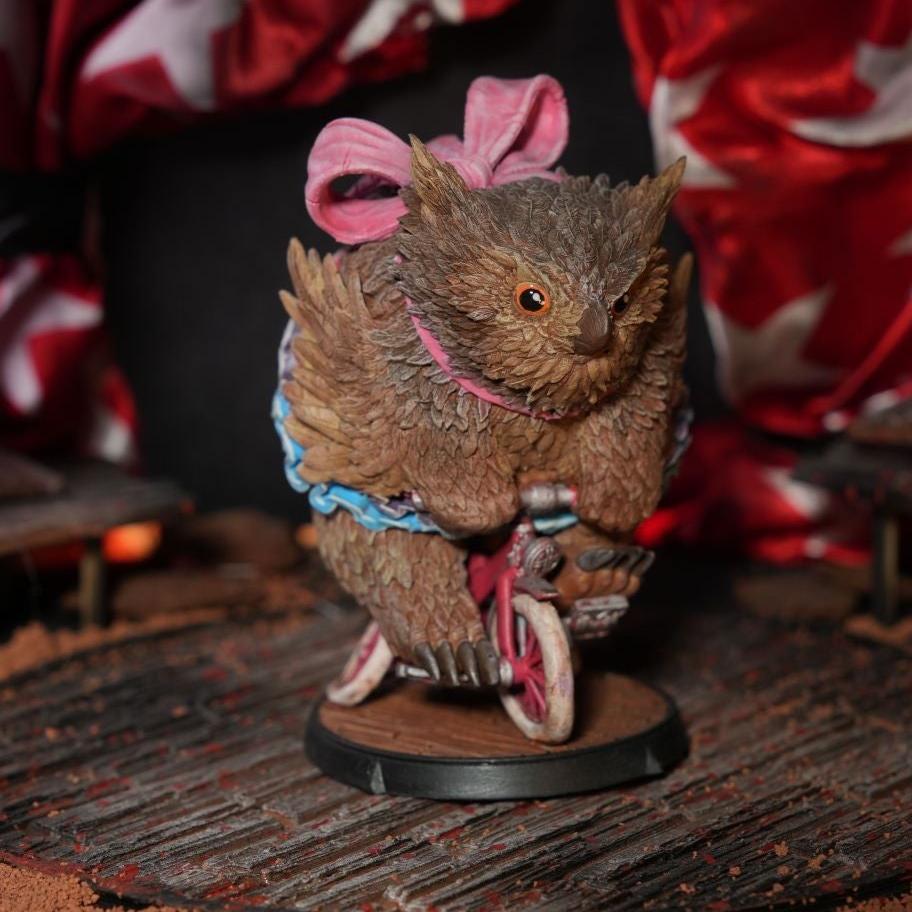 Owlbear miniature riding a unicycle in a circus-themed setting, with detailed feather textures and a dynamic pose, positioned on a wooden stage with draped fabric in the background.