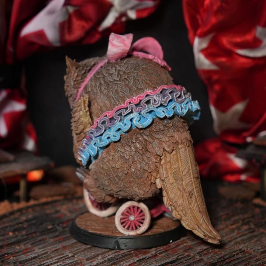 Back view of the Owlbear miniature riding a unicycle, showing detailed feather textures and colorful accessories, set in a circus-themed environment with draped fabric and a wooden stage.